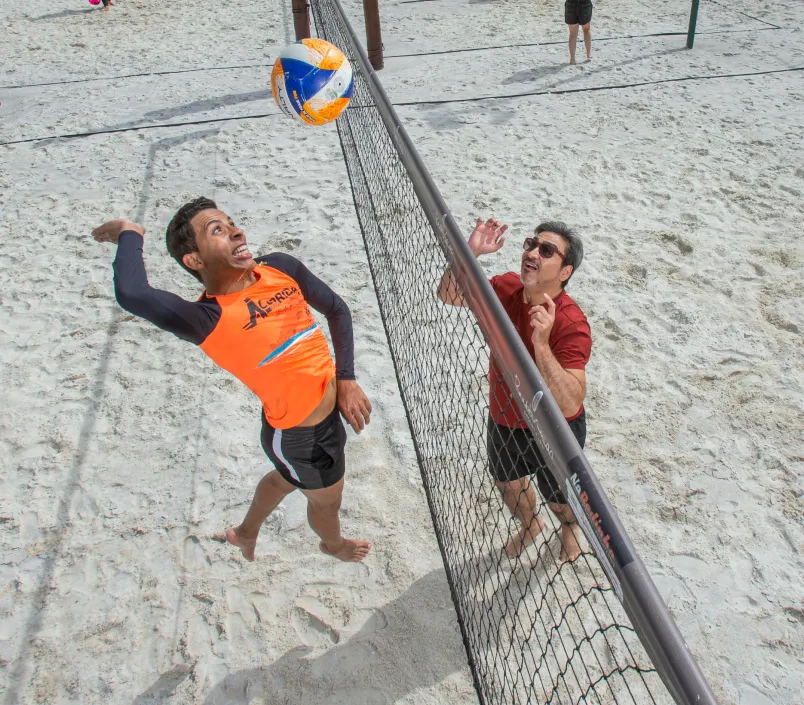 Dois homens estão jogando vôlei na praia e um deles está vestindo uma camisa laranja que diz asics